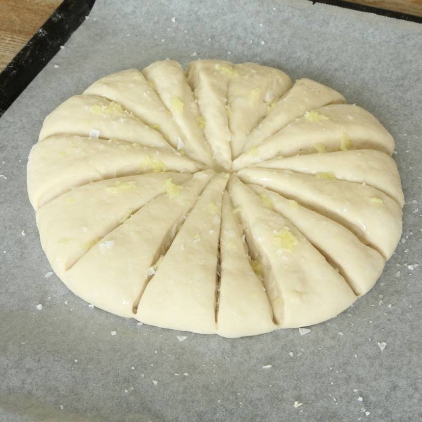 2. Tryck ut degen till en rund kaka, ca 2 cm tjock, på en plåt med bakplåtspapper. Dela degen i sexton bitar med en degskrapa eller kniv. Låt den jäsa under bakduk i ca 30 min. Sätt ugnen på 250 grader. Pressa vitlökslyftan i en skål och blanda med oljan. Pensla blandningen på degen. Strö över flingsalt.